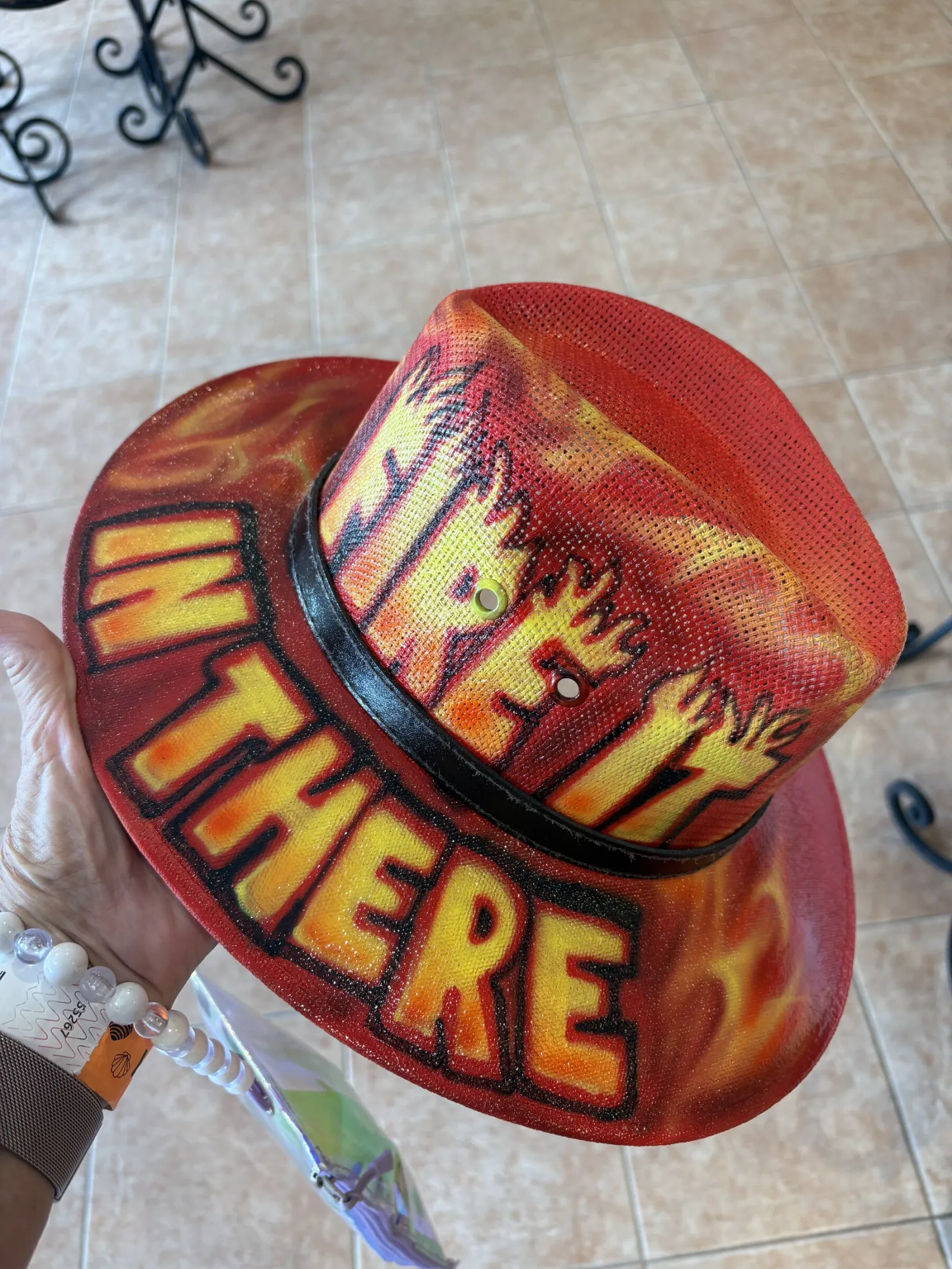 Hand-painted flame hat reading Fire It In There, held up on a tiled terrace.